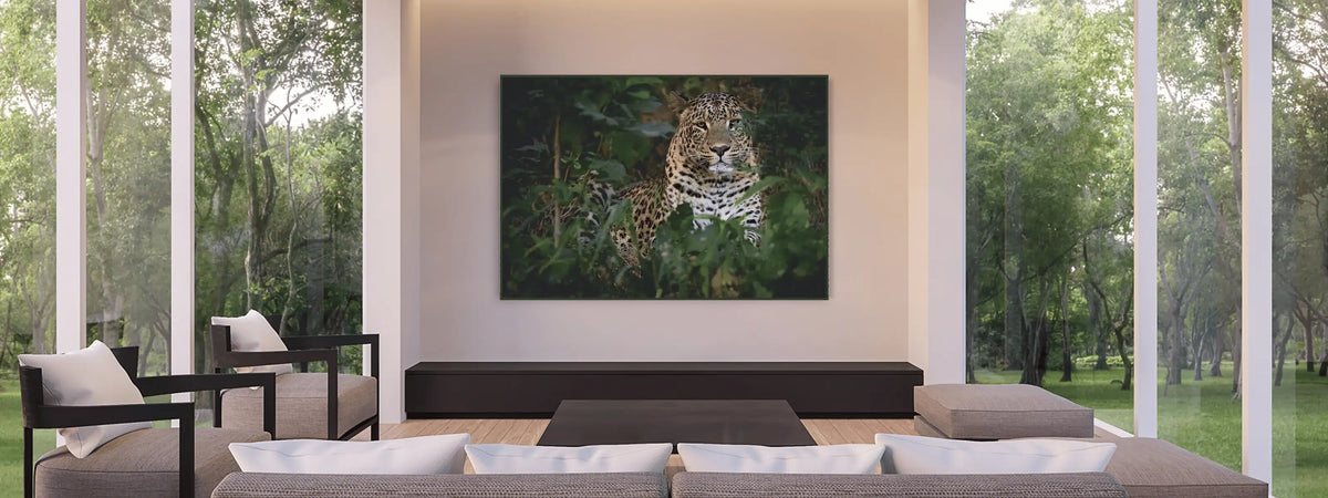 A large framed premium art print depicting a leopard in green foliage, displayed above a dark console table in a modern living room with large windows and outdoor views.